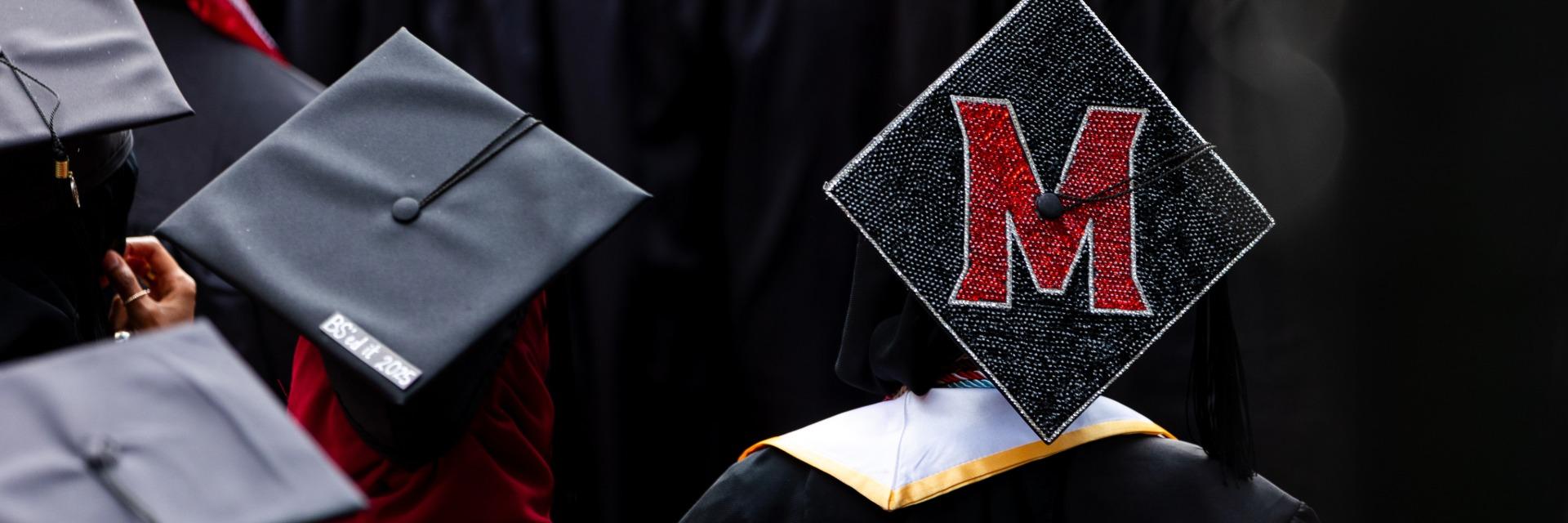 A black graduation cap decorated with a large red and white M in a group of graduates.